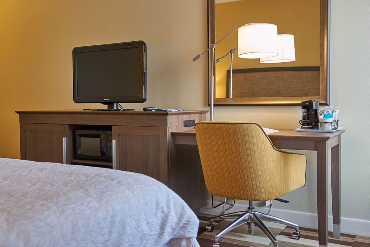 Habitación de hotel con cama, escritorio con computadora, silla, lámpara y cafetera.