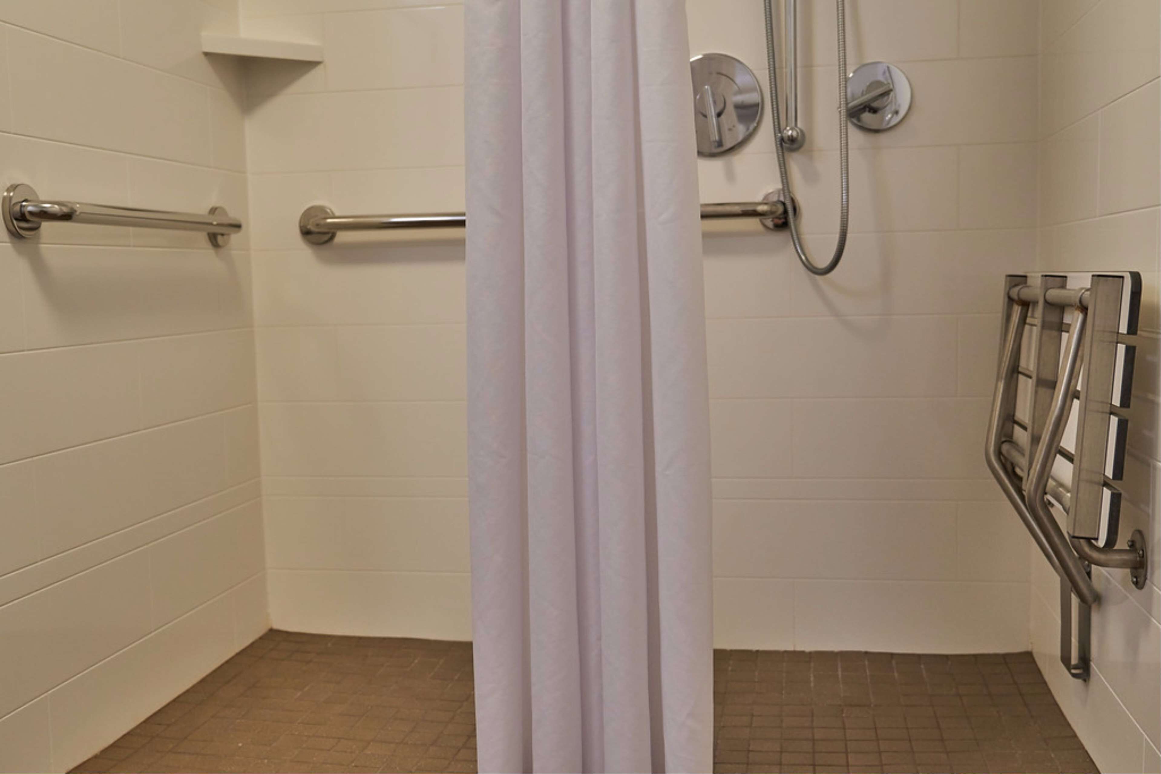 Bagno con pareti piastrellate di bianco, doccia con soffione manuale e maniglione a destra.