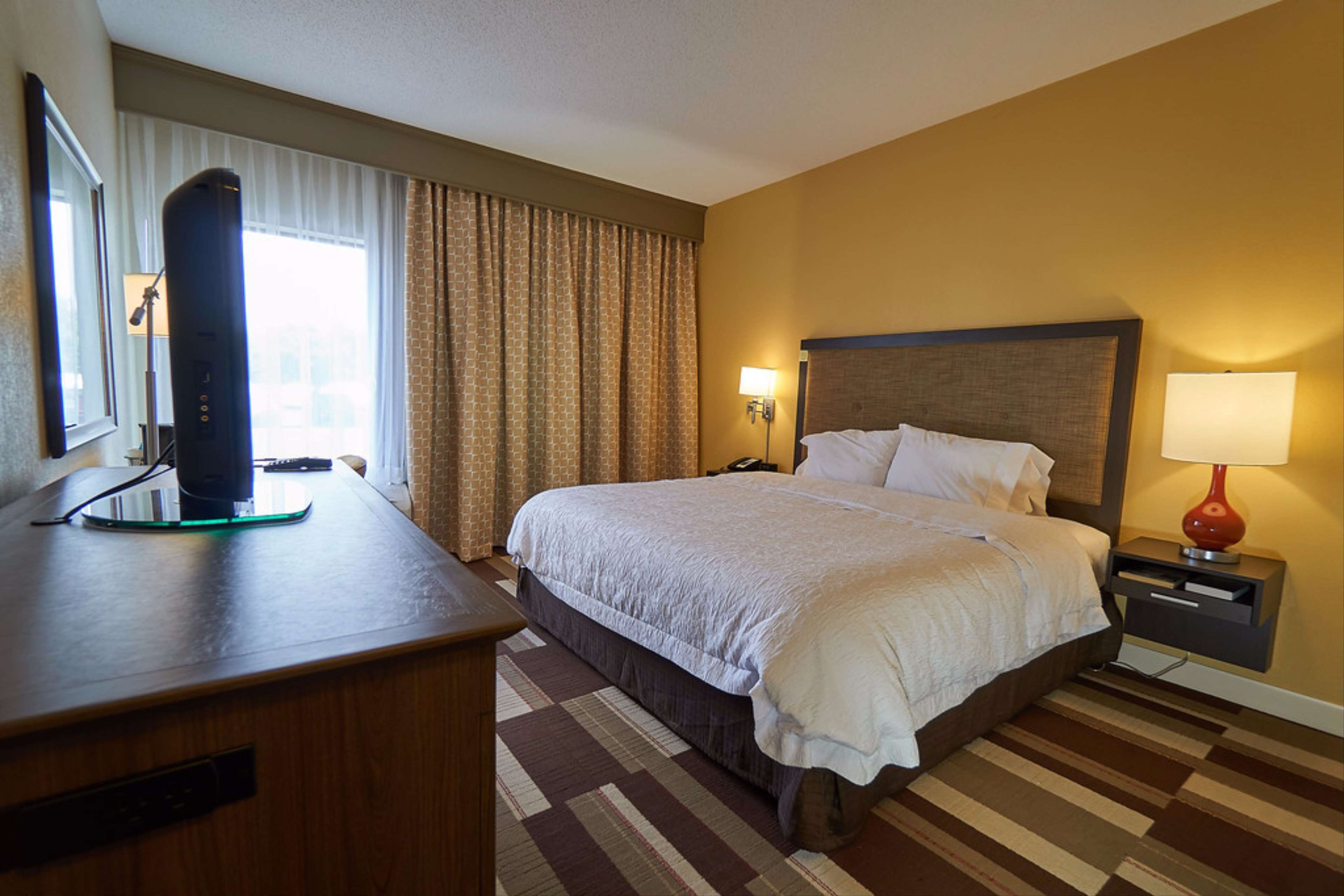 A hotel room with a bed, a desk with a computer, a lamp, and a window with curtains.