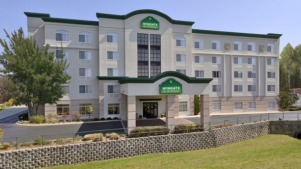 A multi-story hotel with a green and white facade, a central entrance with a sign, and a stone retaining wall in front. - Wingate Inn Chattanooga (Chattanooga)
