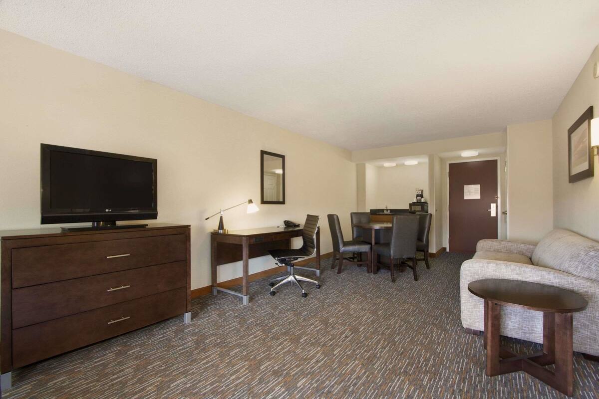 Quarto de hotel com televisão de tela plana, uma escrivaninha com cadeira, um sofá e uma pequena mesa.