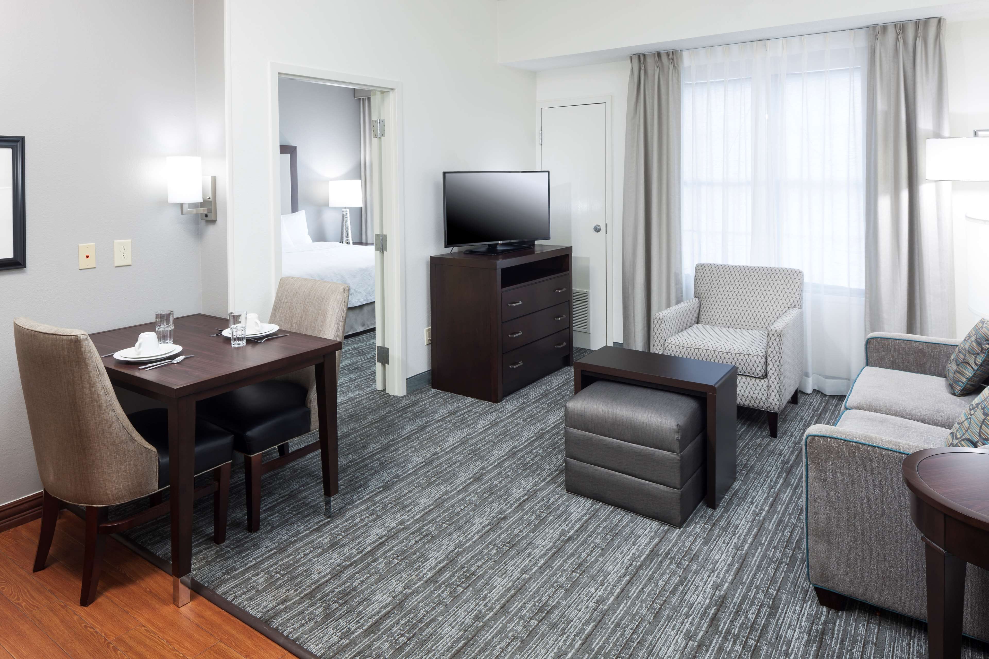 A hotel room with a dining table, chairs, a TV on a cabinet, a sofa, and a patterned armchair.