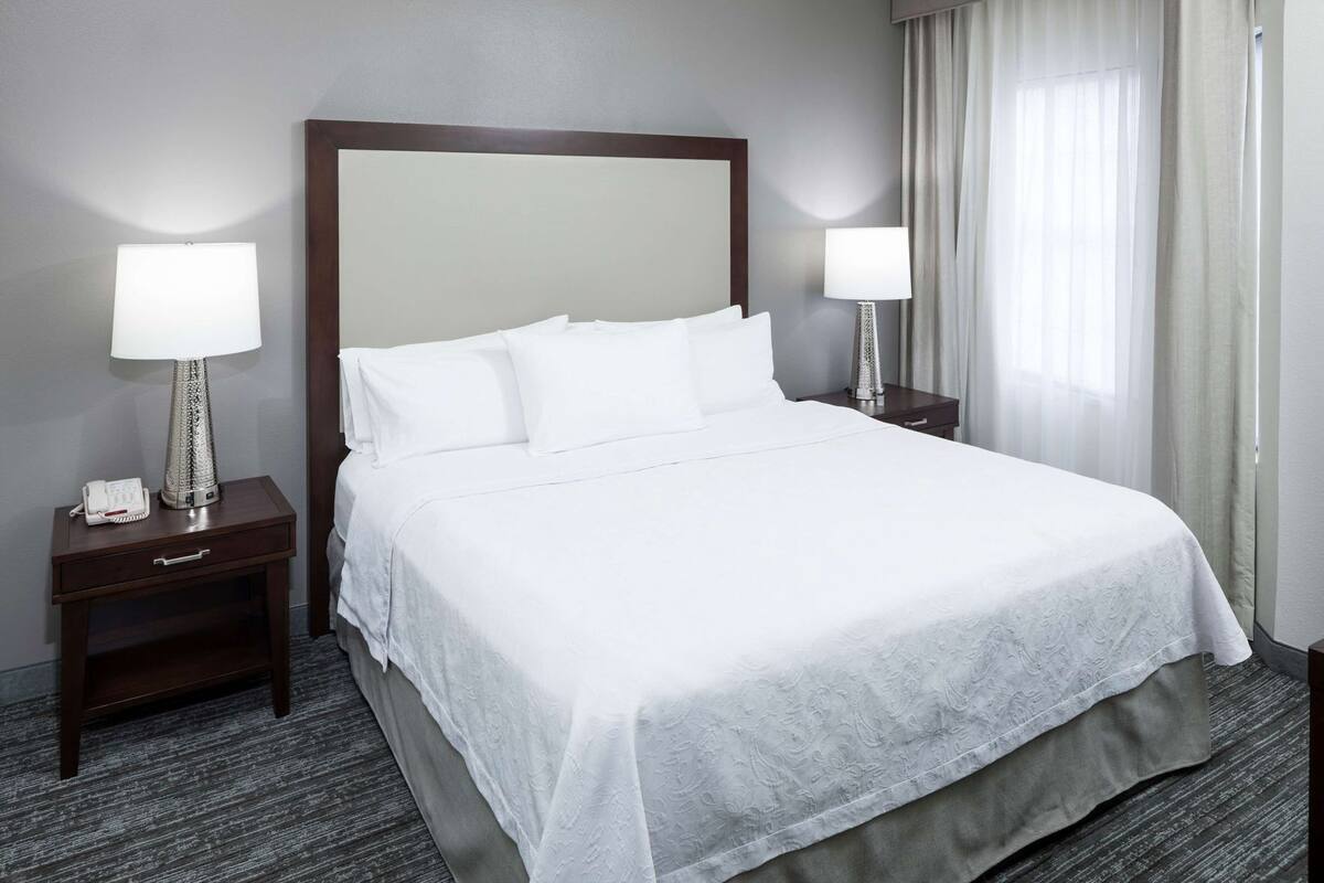 Een net opgemaakt bed met witte lakens, twee bedlampjes, een telefoon op het nachtkastje links, en een houten hoofdbord.