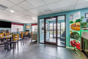A restaurant with a modern interior, featuring a large dining area with tables and chairs, a kitchen area with a TV, and a glass door leading outside.