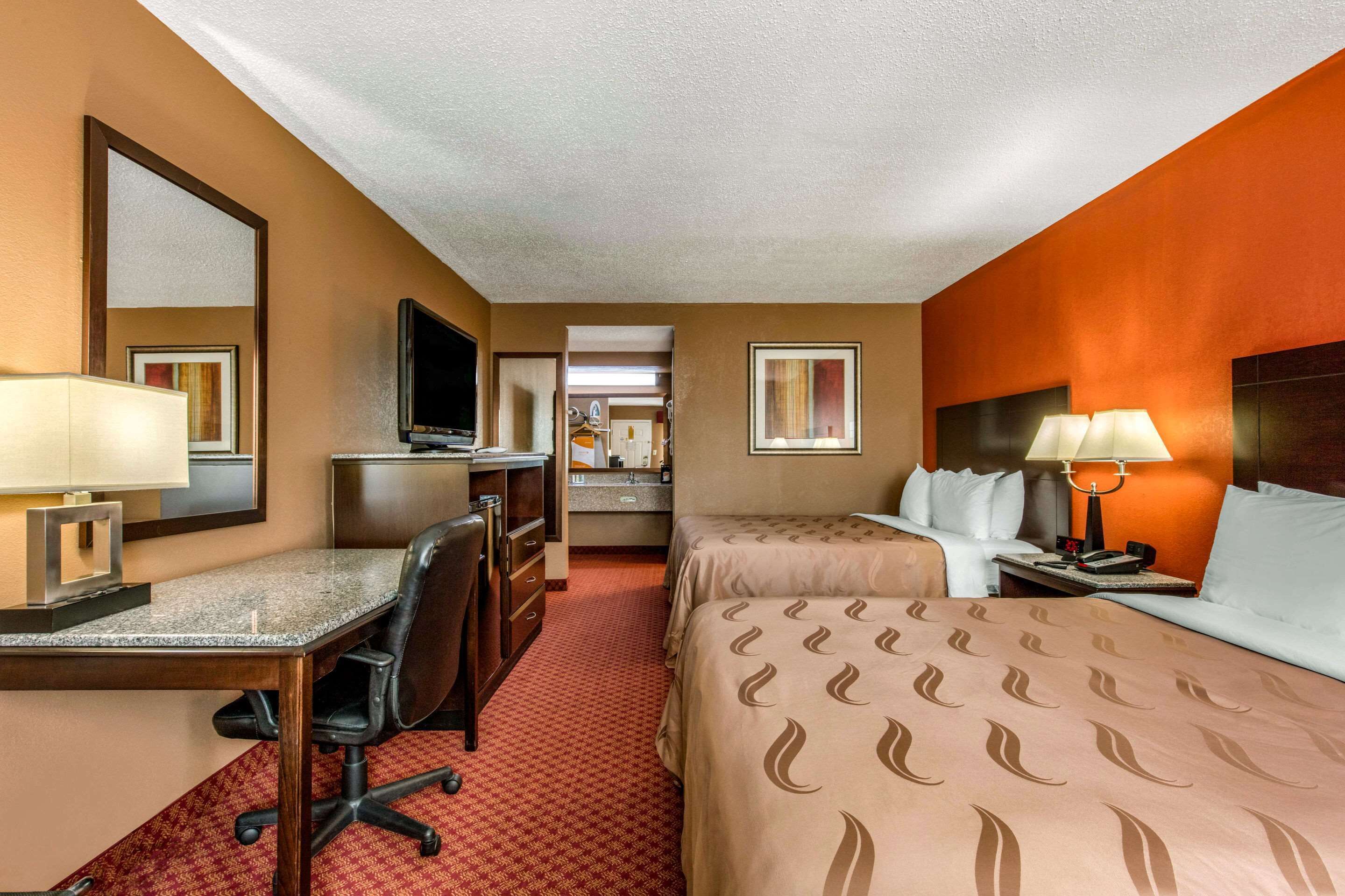 Une chambre d’hôtel équipée d’un bureau, d’une chaise, de deux lits, d’une télévision et d’une salle de bain visible à travers une porte ouverte.