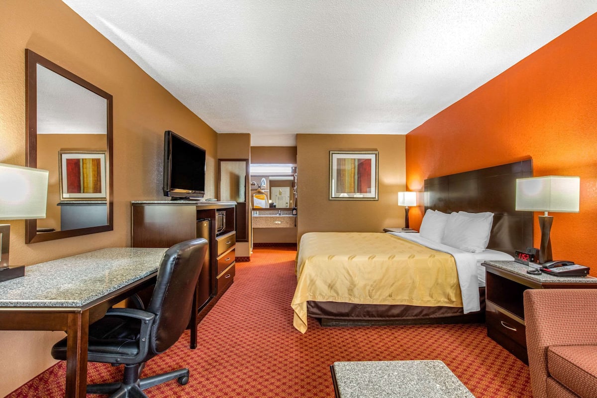 A hotel room with a bed, desk, chair, television, and a bathroom visible in the background.