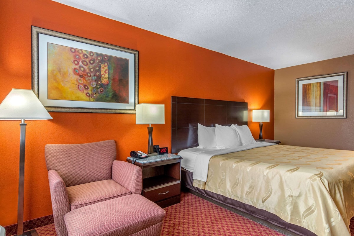 A hotel room with an orange wall, a large abstract painting, a bed with white linens, two bedside lamps, a red patterned armchair, and a nightstand with a phone and clock.