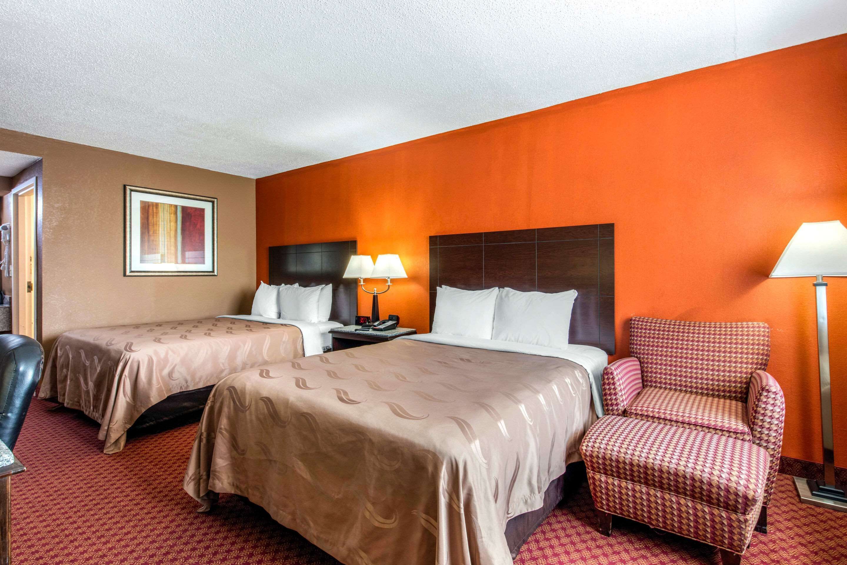 A hotel room with two beds, a red armchair, and a nightstand with lamps.