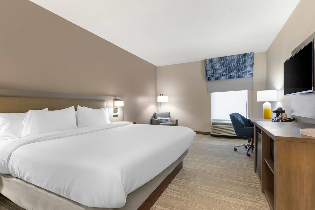 A hotel room with a large bed, a desk with a chair, a flat-screen TV, and a window with a view of the city.