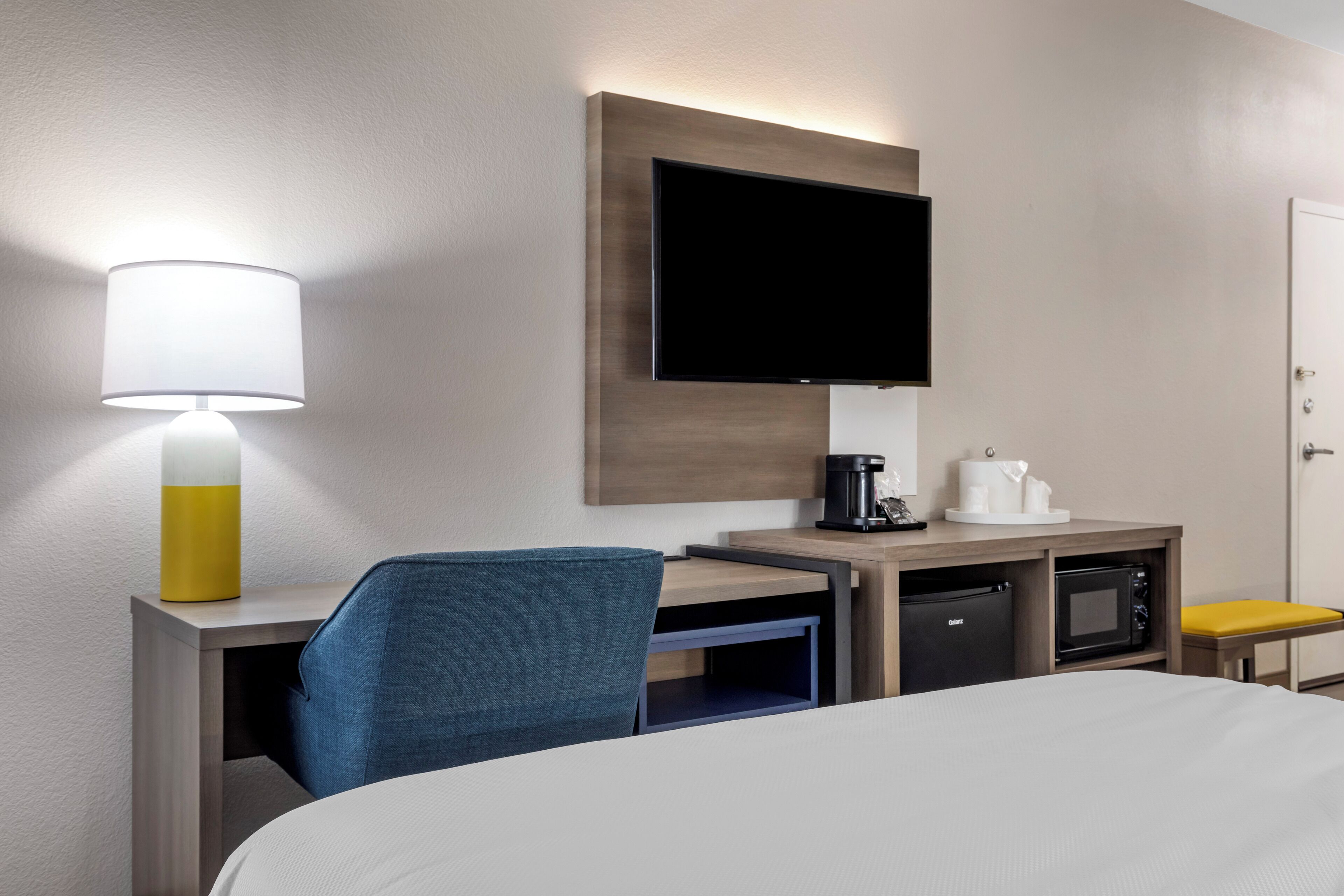 Habitación de hotel con cama, escritorio con lámpara, televisor, microondas y cafetera.