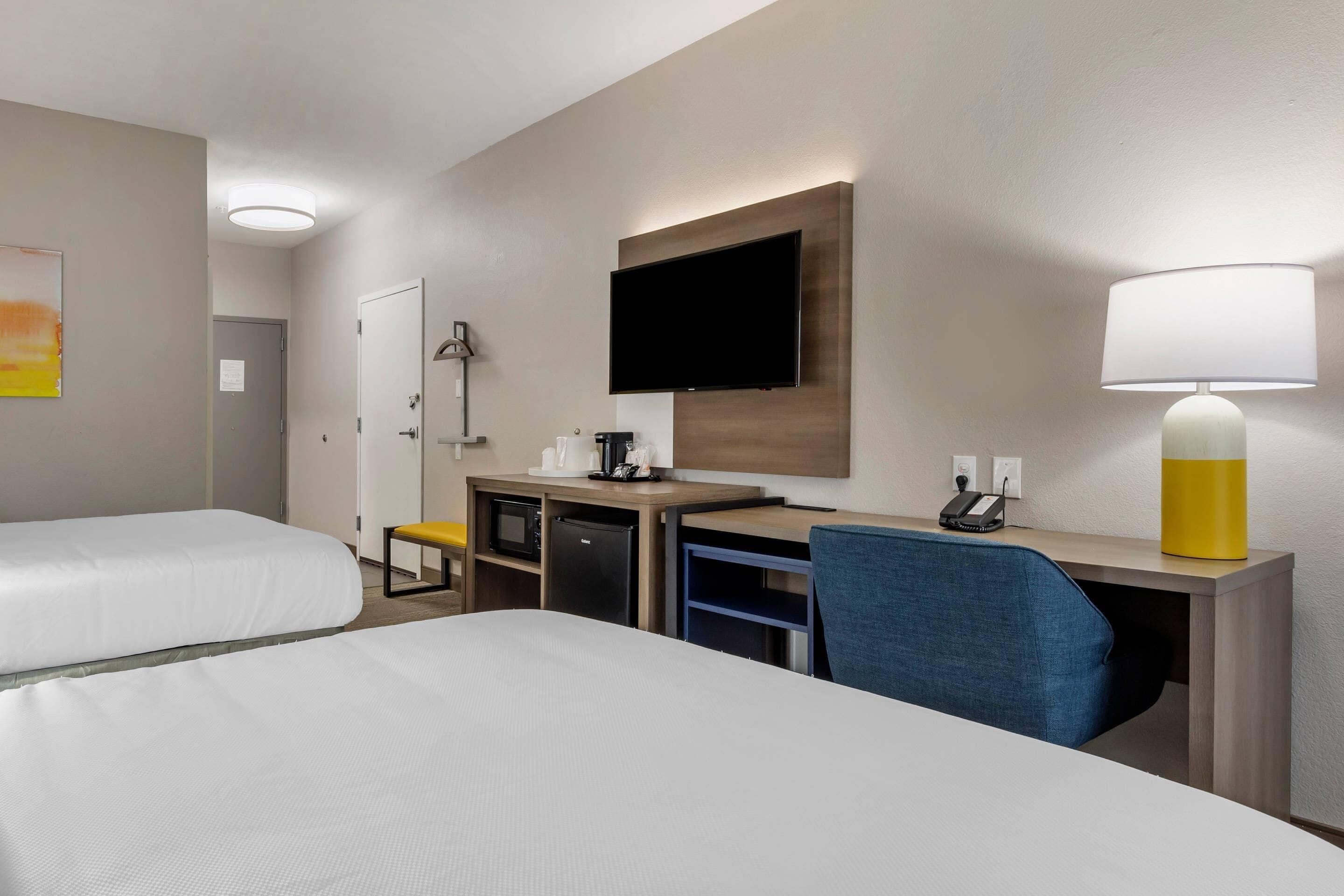 A hotel room with two beds, a desk with a computer, a TV, a lamp, and a small kitchenette.