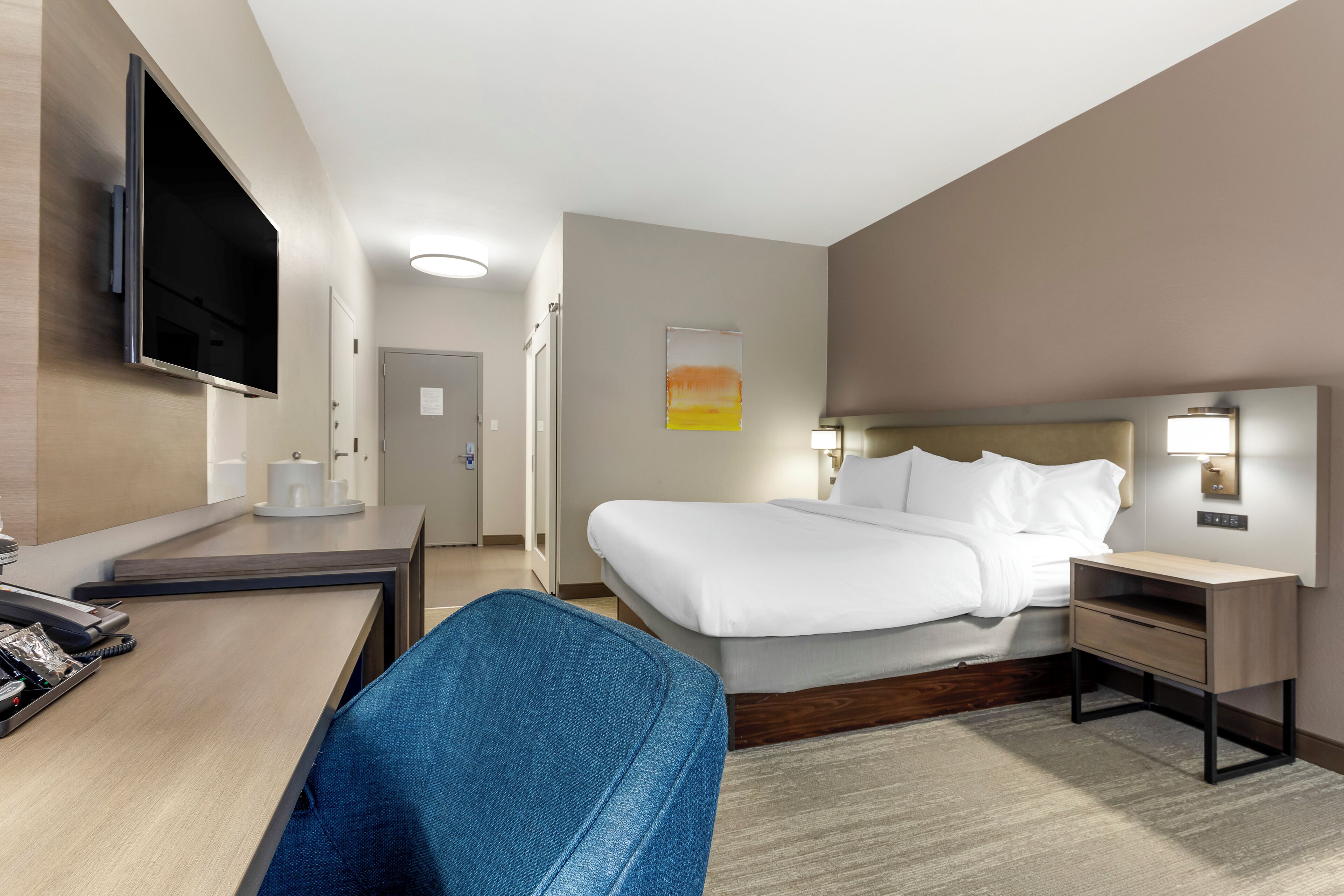 Habitación de hotel con cama, escritorio, silla, televisor y mesita de noche con lámpara.