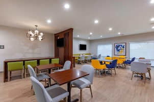 Un comedor moderno con mesas y sillas de madera, una zona de estar central con respaldo alto, y un televisor de pantalla plana montado en la pared.