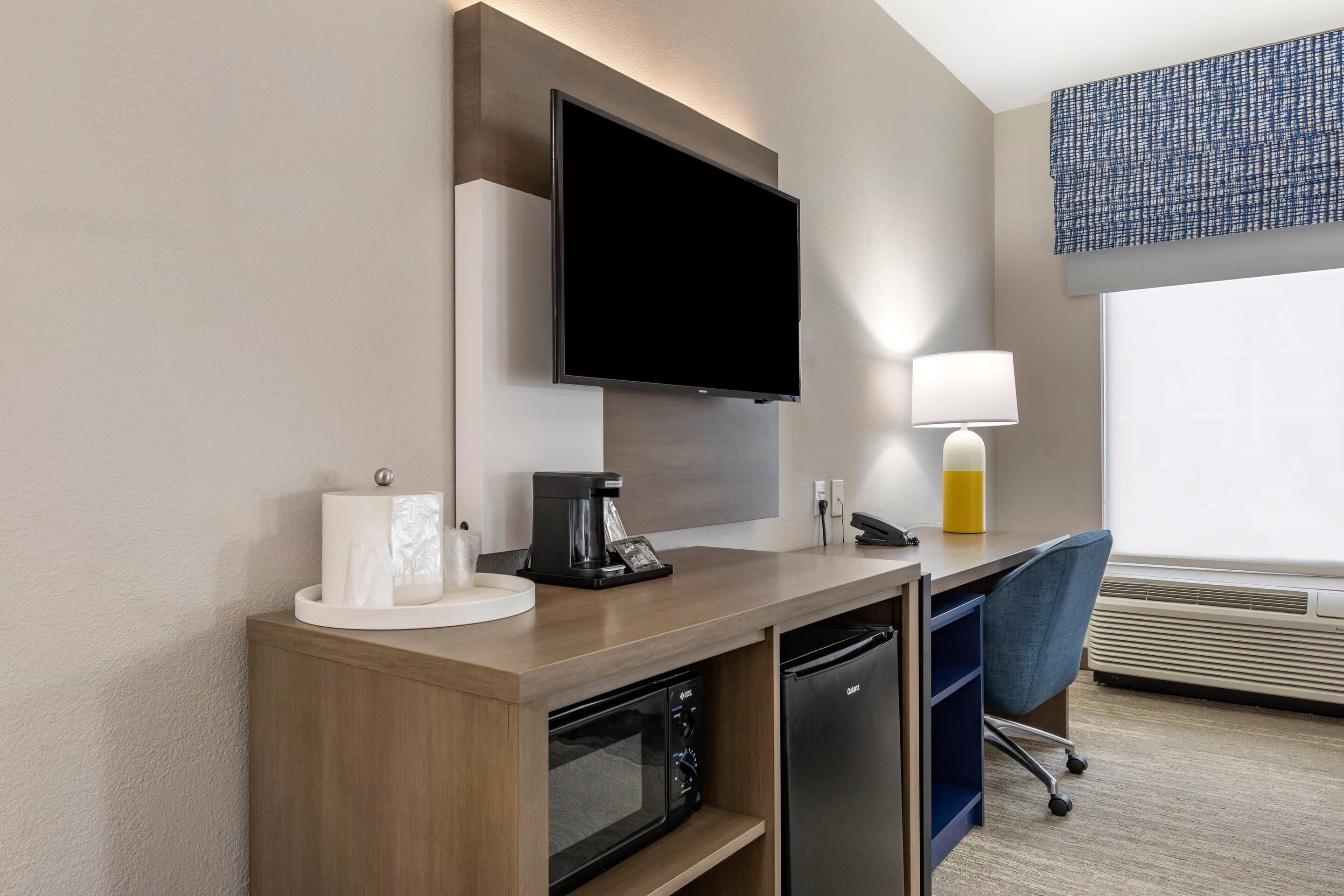 Habitación de hotel con televisión de pantalla plana montada en la pared, cafetera, lámpara, escritorio con silla, microondas y refrigerador.