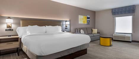 A modern hotel room with a large bed, bedside tables, a sofa, a small round table, and a window with an air conditioning unit.