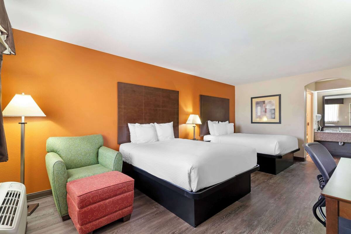 Ein Hotelzimmer mit zwei Betten, einem grünen Sessel, einem roten Hocker, einem Schreibtisch mit Stuhl und einem großen Spiegel.
