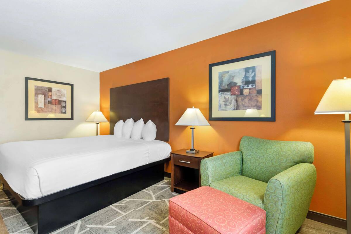 Ein Hotelzimmer mit einem Bett, einem grünen Sessel, einem roten Hocker, zwei Wandlampen und zwei gerahmten Bildern an der Wand.