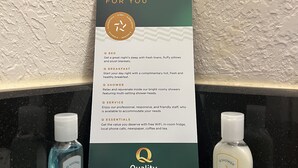 A hotel bathroom with a sign detailing amenities like a bed, breakfast, shower, service, and essentials. There are two bottles, one of shampoo and one of lotion, on the counter. - Quality Inn & Suites Houston Northwest - Cypress (Houston)