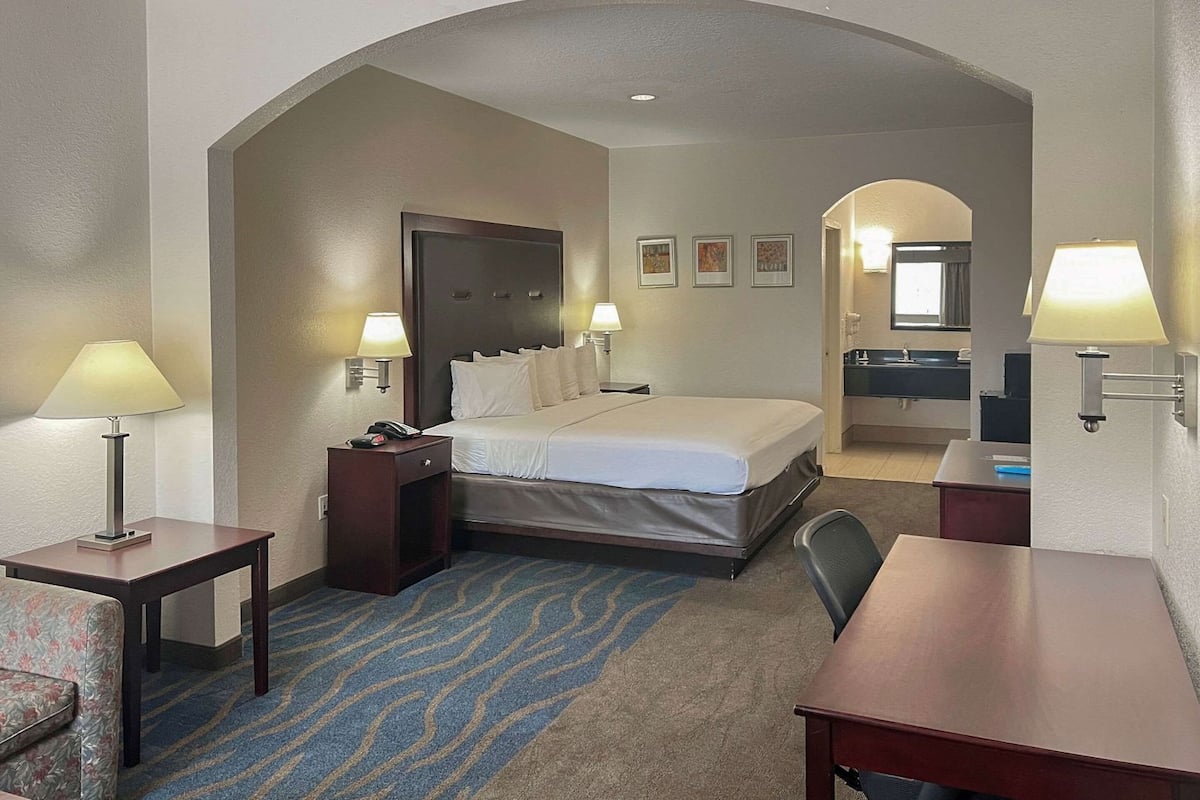 A hotel room with a bed, two bedside tables, a desk, a chair, and a bathroom visible through an archway.