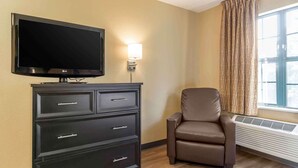 Une chambre d’hôtel avec un téléviseur à écran plat posé sur un meuble noir, un fauteuil en cuir marron et une fenêtre avec des rideaux.