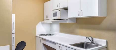 A compact kitchenette with white cabinets, a microwave, a sink, and a chair.