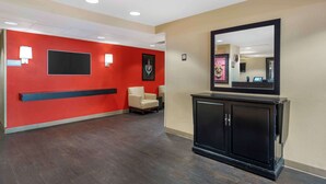 Un hall d’hôtel avec un mur rouge, un comptoir d’accueil noir, un fauteuil beige et un grand miroir.