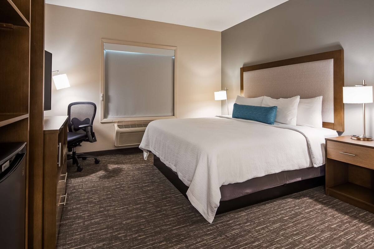 A hotel room with a large bed, a desk with a chair, a wardrobe, and a window with blinds.