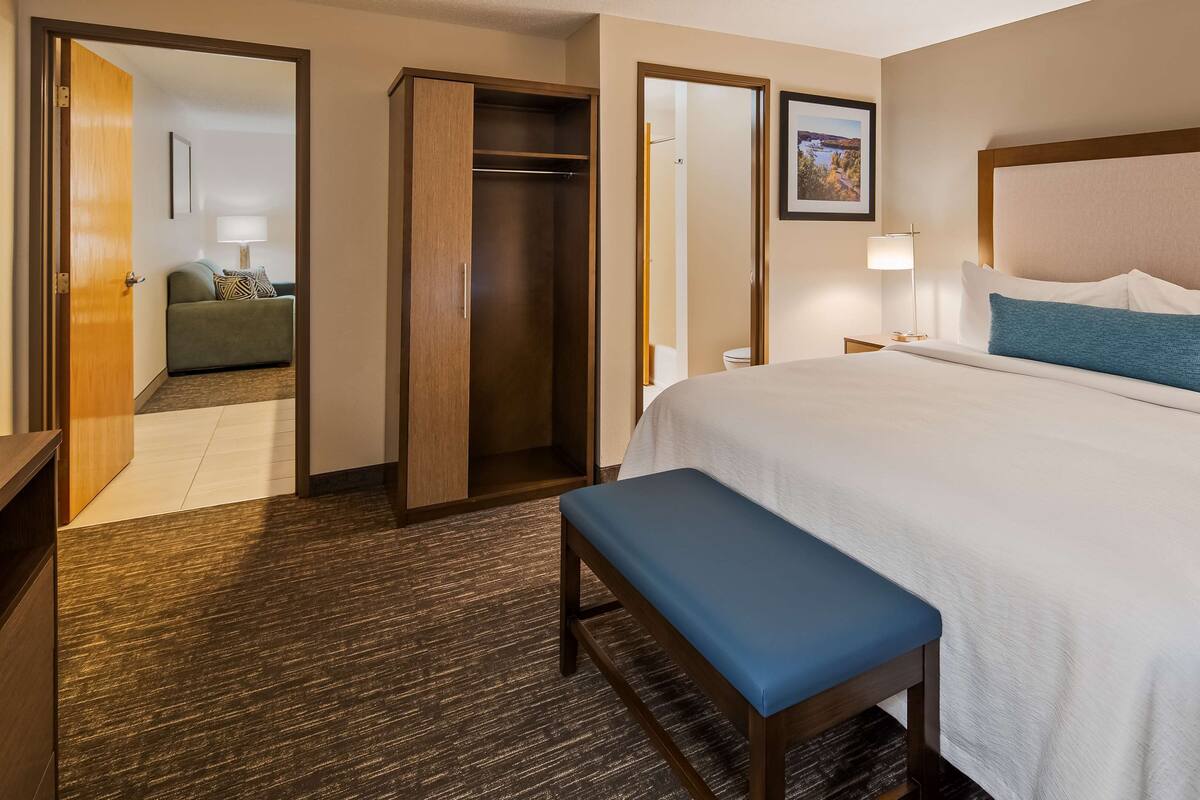 A hotel room with a large bed, a bench, a wardrobe, and a bathroom visible through an open door.