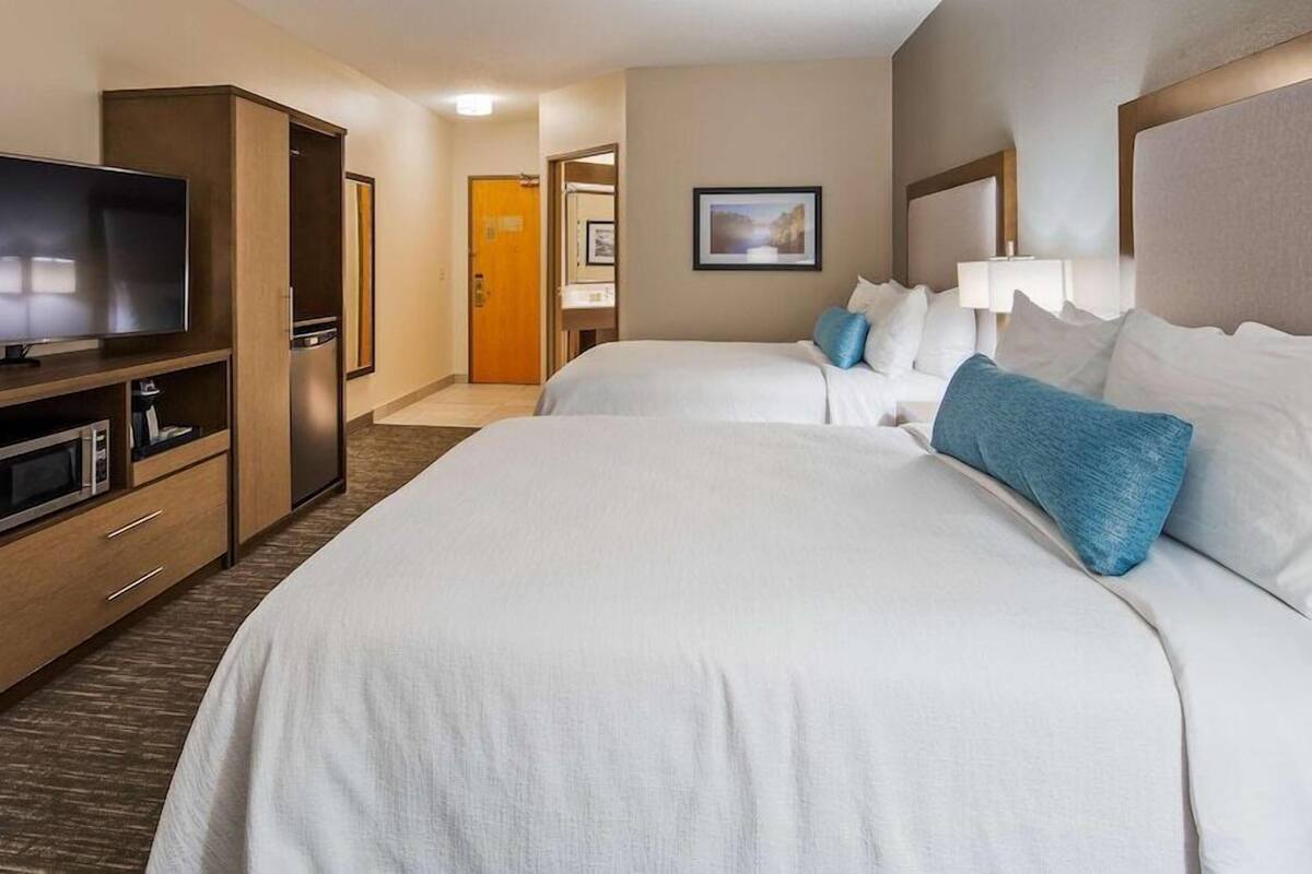 A hotel room with a TV, a minibar, a bed with pillows, and a door leading to another room.