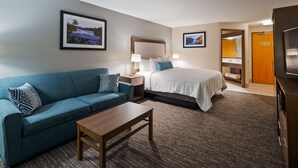 A hotel room with a blue sofa, a wooden coffee table, a bed with white bedding, two bedside tables with lamps, and two framed pictures on the walls. - Best Western Plus Hudson I-94 (Hudson)