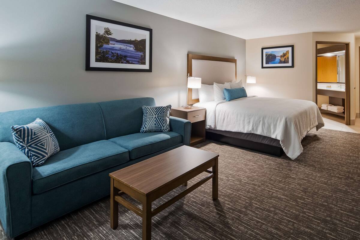 A hotel room with a blue sofa, a wooden coffee table, a bed with white bedding, two bedside tables with lamps, and two framed pictures on the walls.