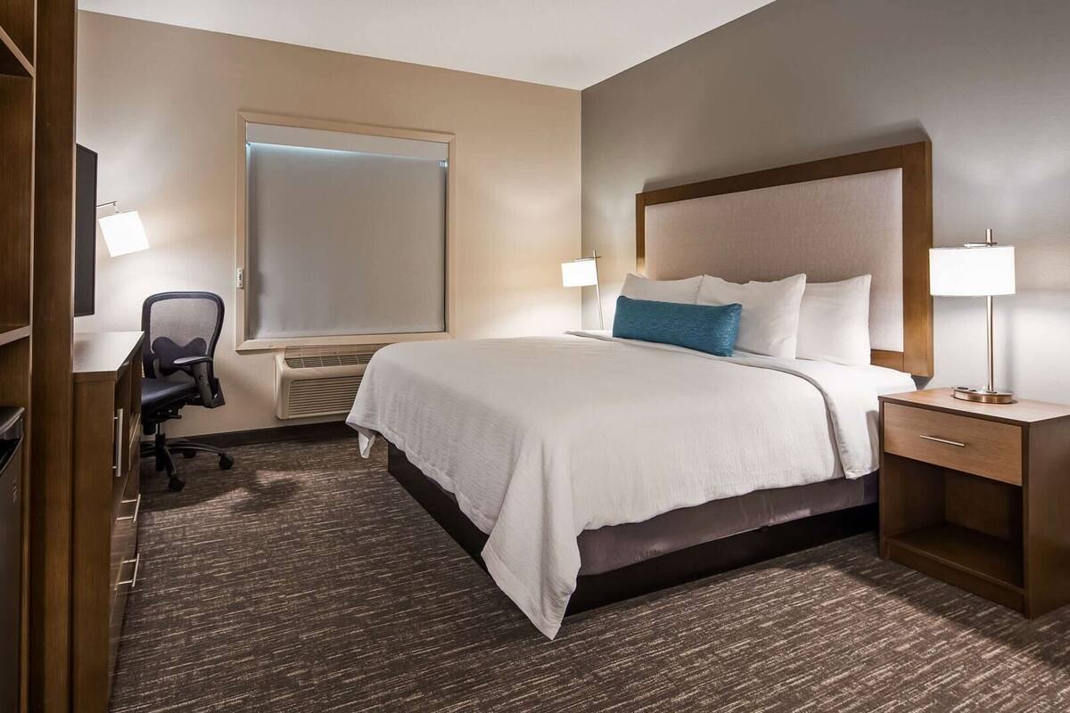 A hotel room with a bed, bedside tables, a desk with a chair, a wardrobe, and a window.