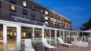A poolside area with lounge chairs and a fire pit, surrounded by glass walls and a building in the background.