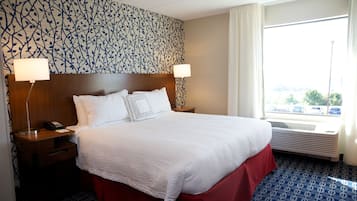A hotel room with a large bed, two bedside tables with lamps, a patterned wall, and a window with a view.