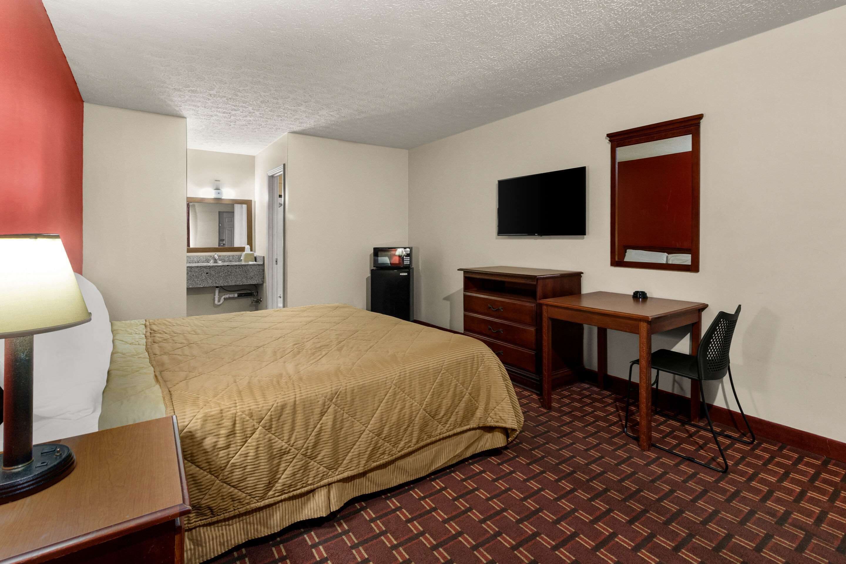A hotel room with a bed, a desk, a chair, a television, and a bathroom visible in the background.