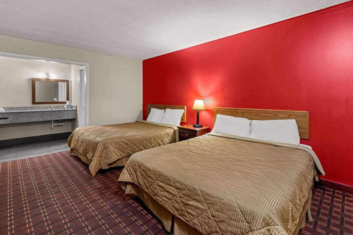 A hotel room with two beds, a red wall, a wooden headboard, a nightstand with a lamp, and a bathroom with a mirror and sink.