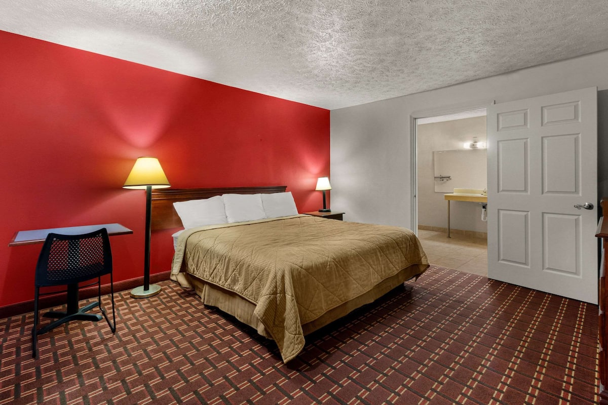 A hotel room with a bed, two bedside tables with lamps, a chair, a red wall, a patterned carpet, and a bathroom visible through an open door.