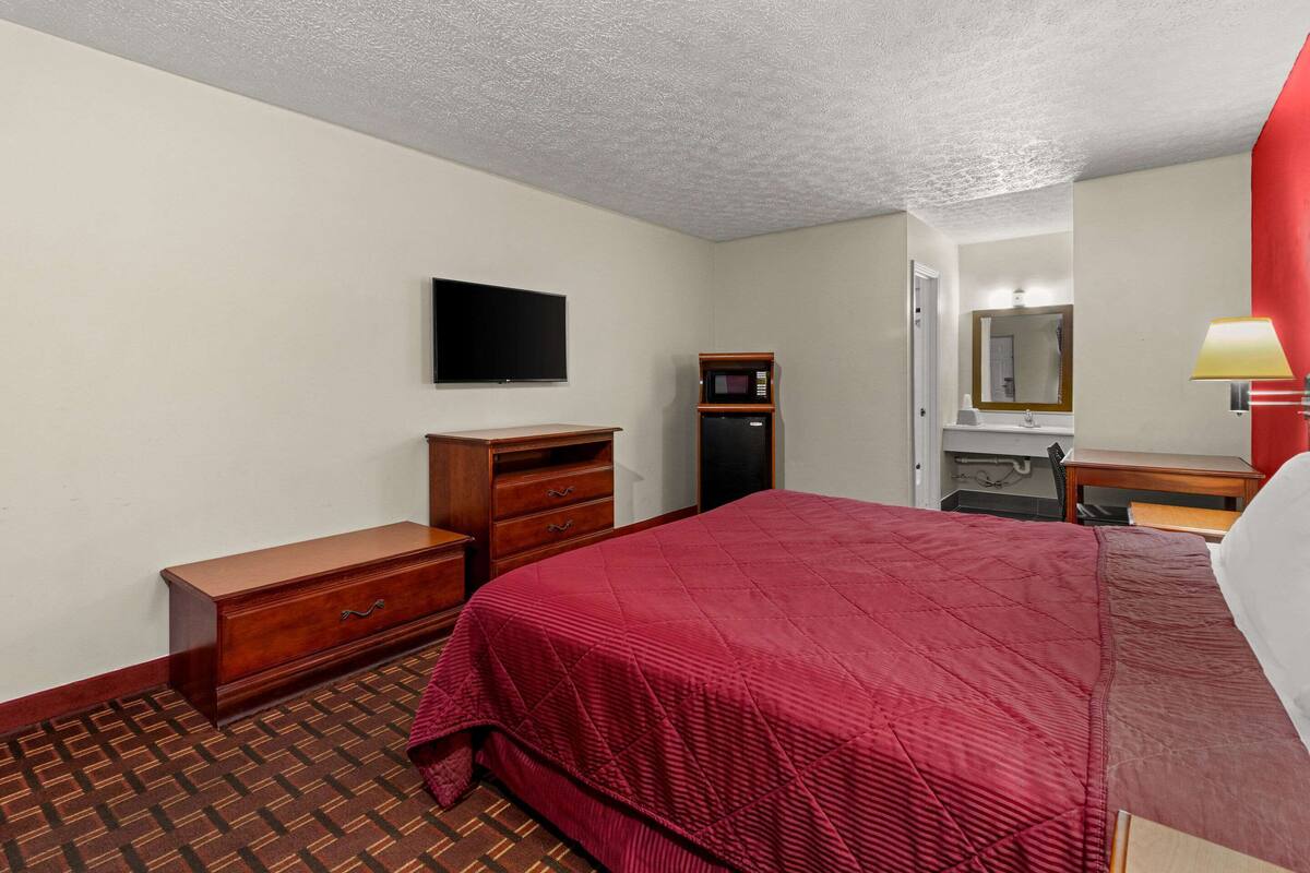 A hotel room with a bed, a wooden dresser, a microwave, a television, and a mirror.