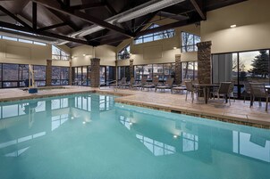 A large indoor swimming pool with clear blue water, surrounded by lounge chairs and tables, large windows offering views of the outside.