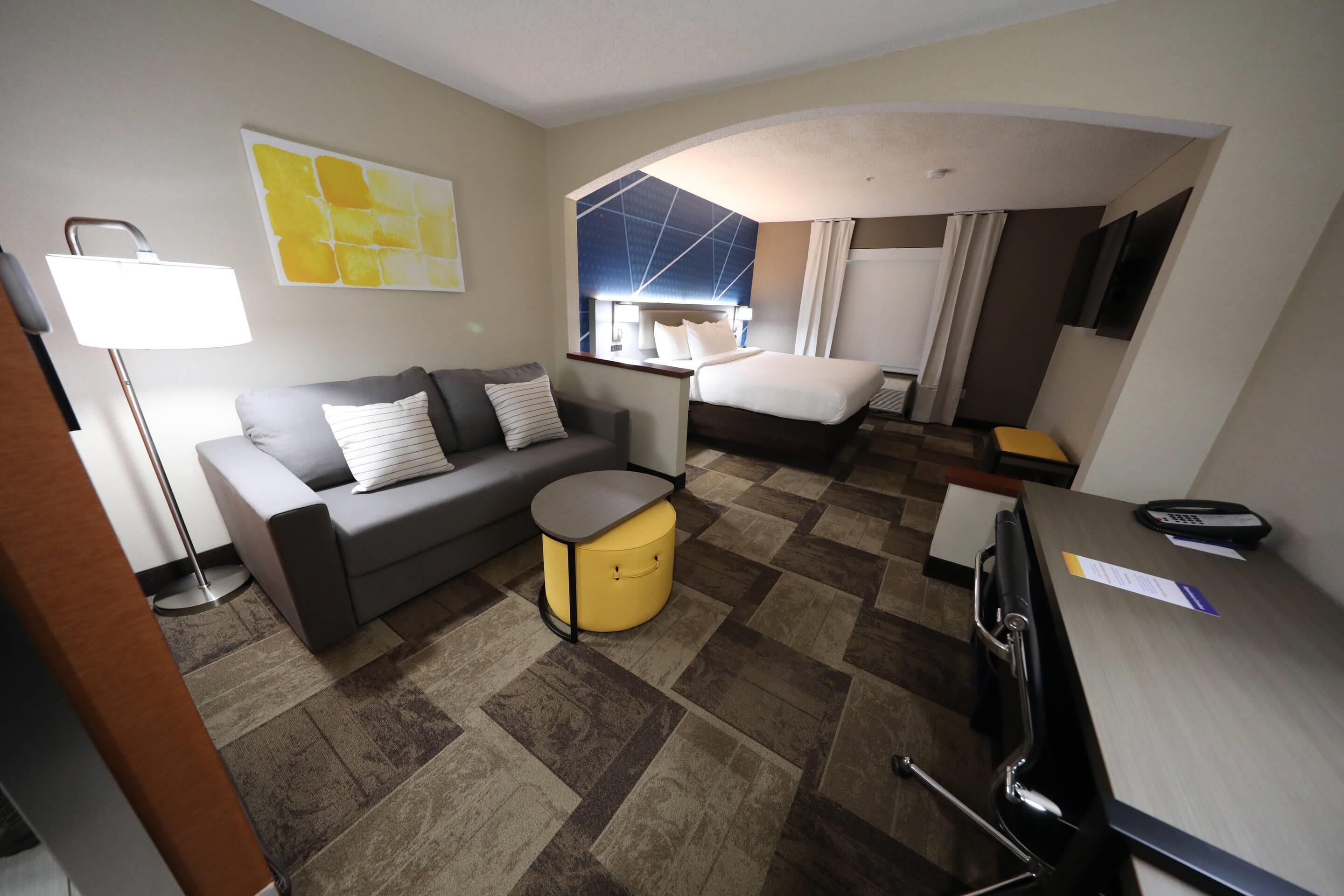 Una habitación de hotel con sofá, un reposapiés amarillo pequeño, una cama, un escritorio con teléfono y una lámpara.