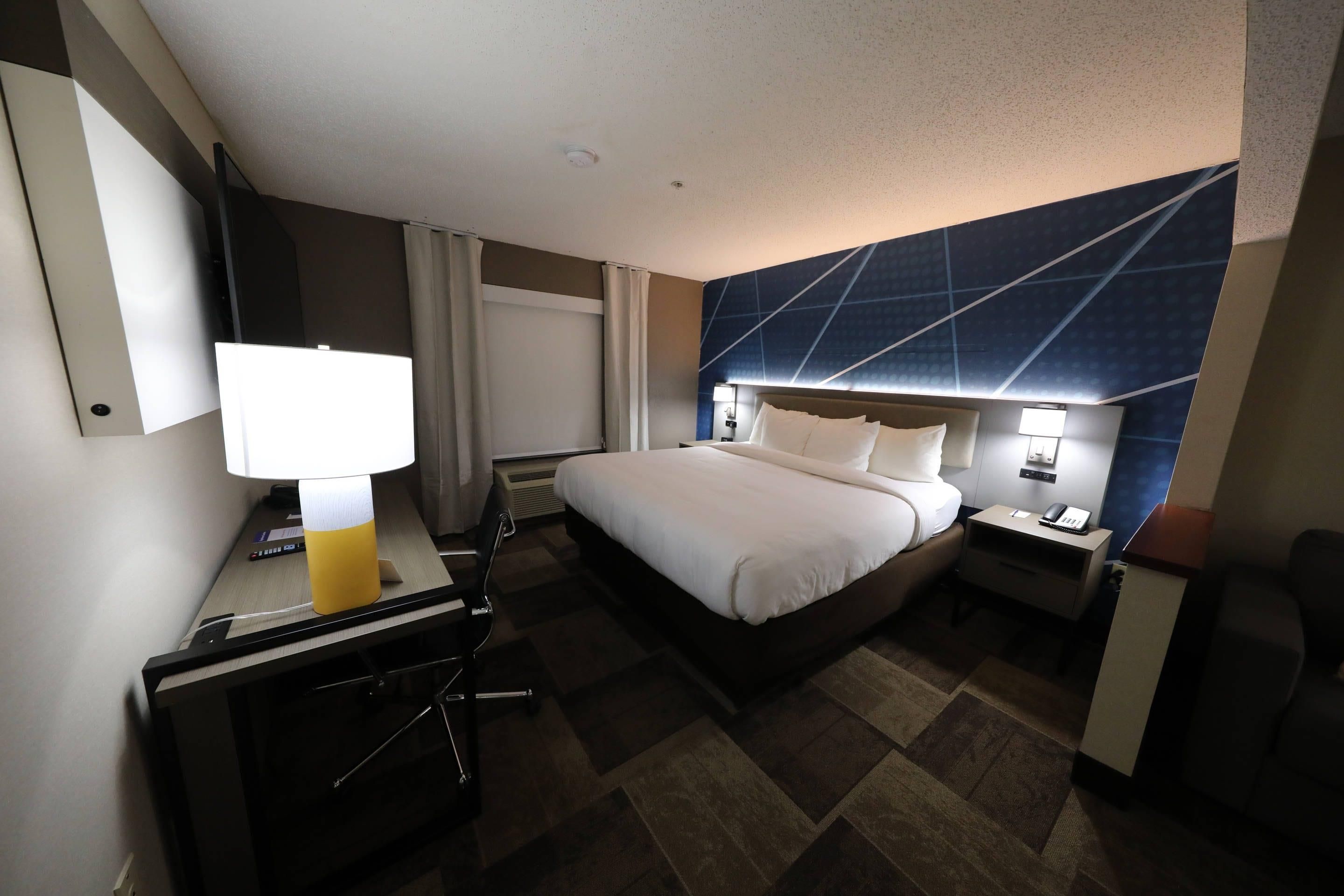 A hotel room with a bed, bedside tables, a desk with a lamp, a chair, and a large window with curtains.
