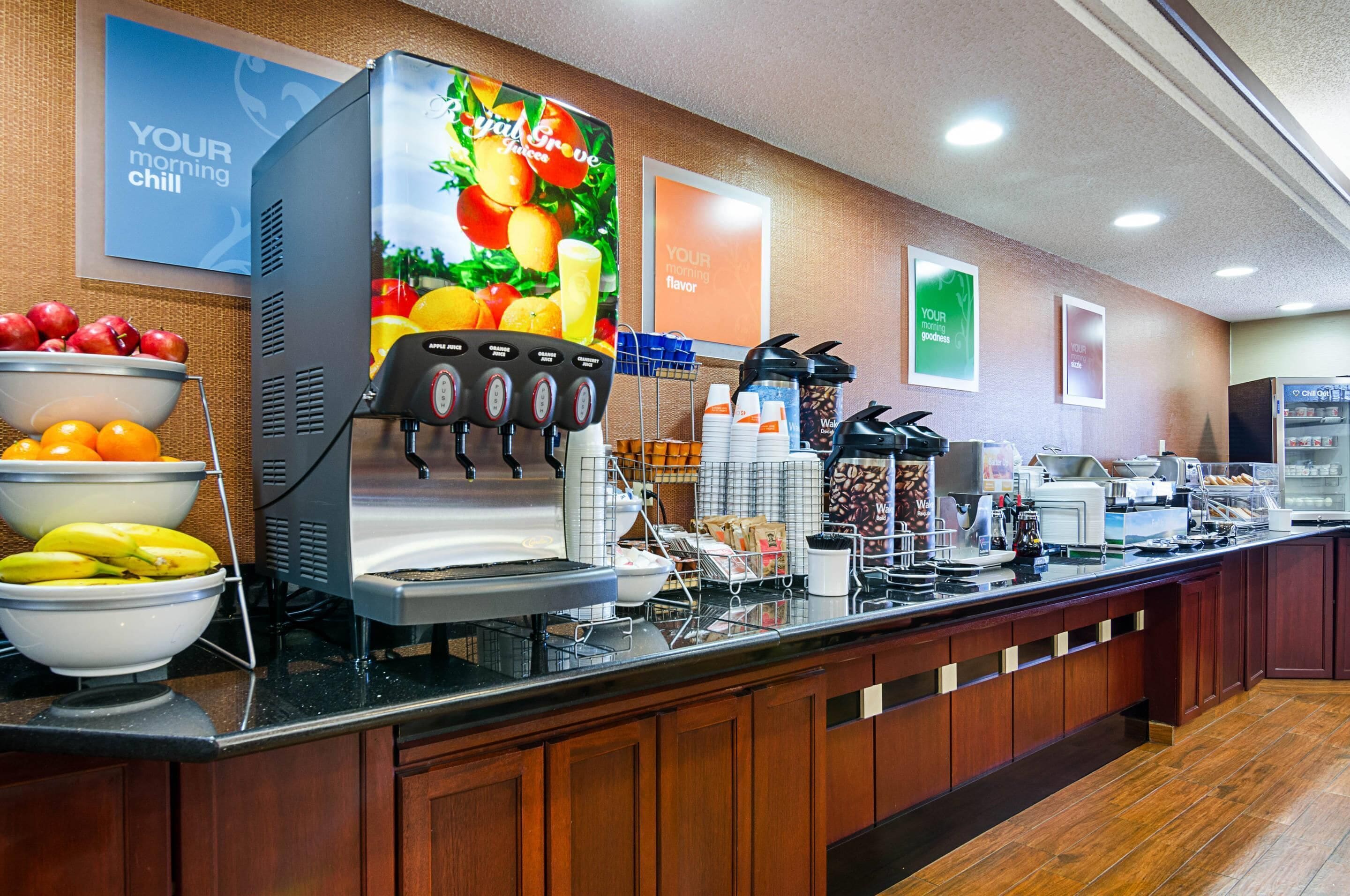 A self-service breakfast bar with fruit bowls, a coffee machine, and a juice dispenser.