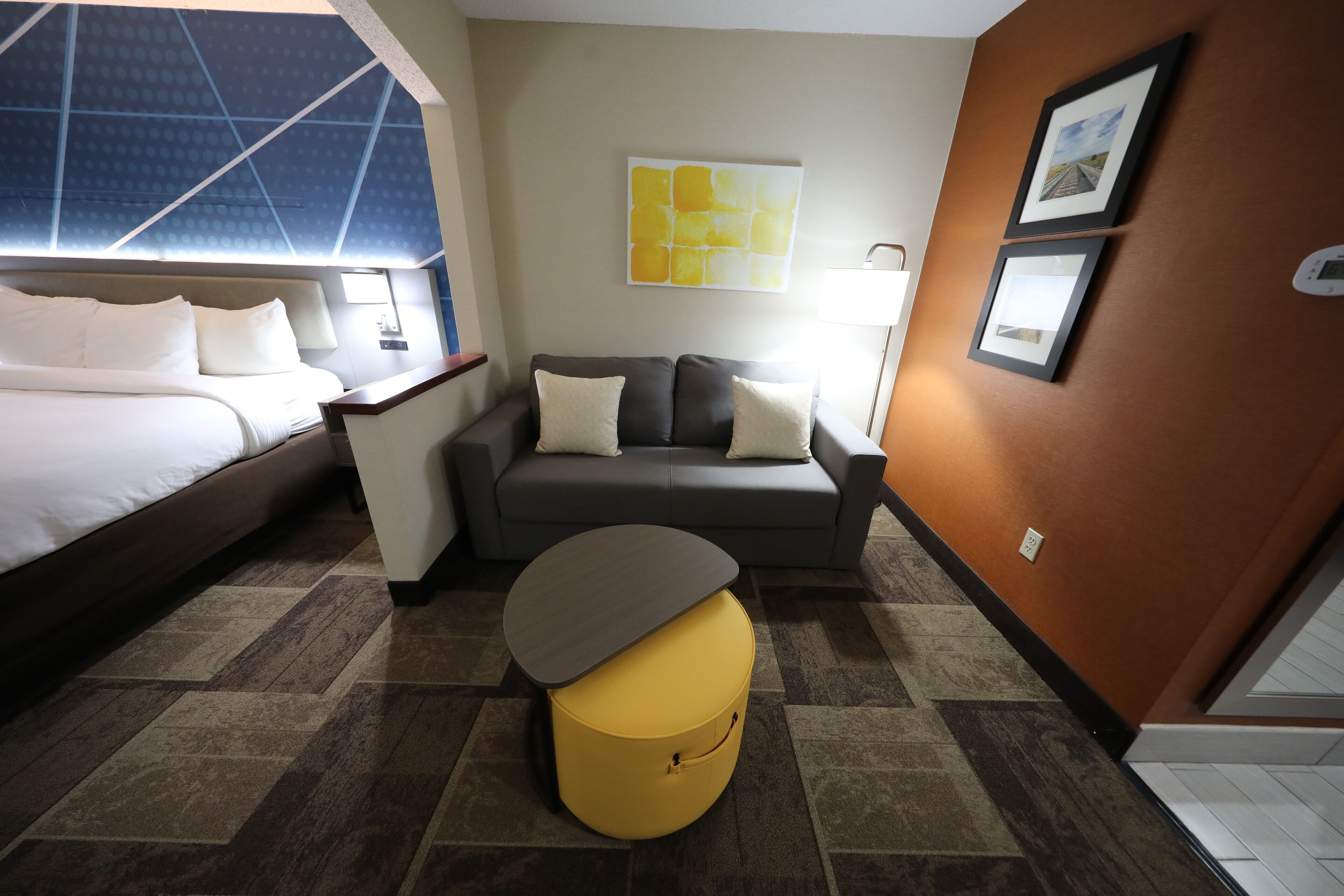 A hotel room with a bed, a sofa, a small table, and a yellow ottoman.