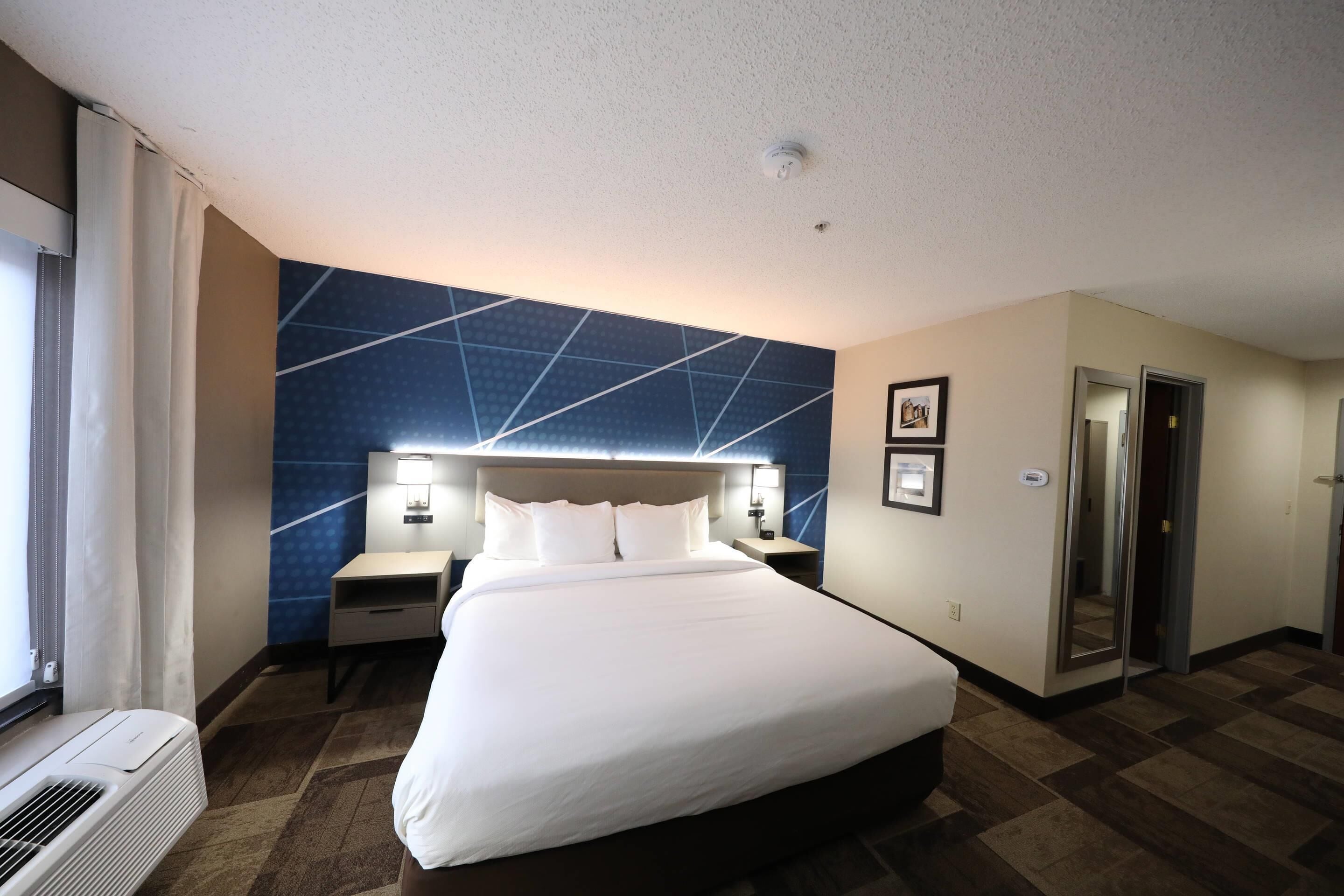 A hotel room with a large bed, a nightstand, a wall-mounted lamp, a window with curtains, and a door.