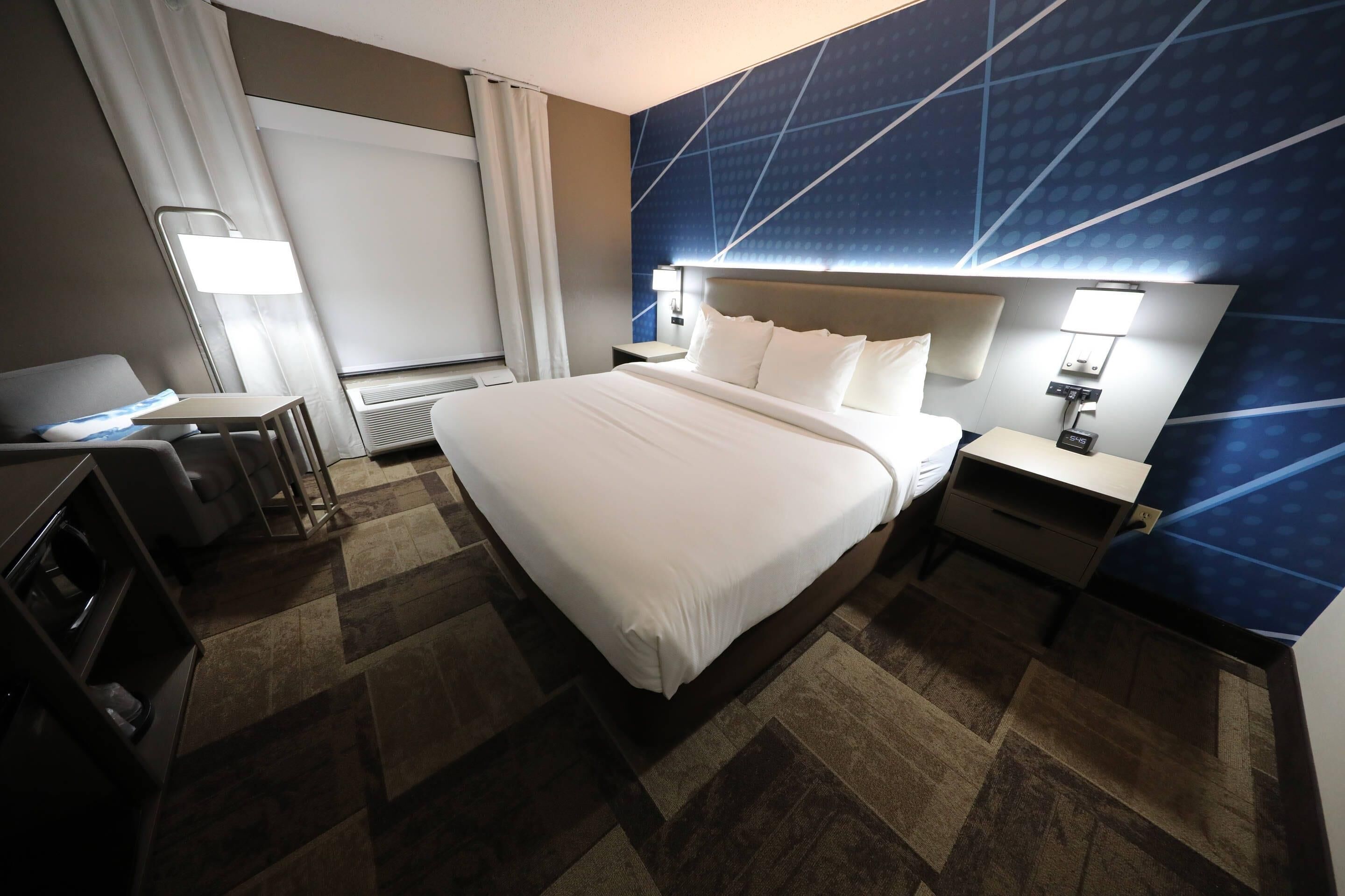 A hotel room with a large bed, a desk, a chair, a nightstand, a lamp, and a window with curtains.