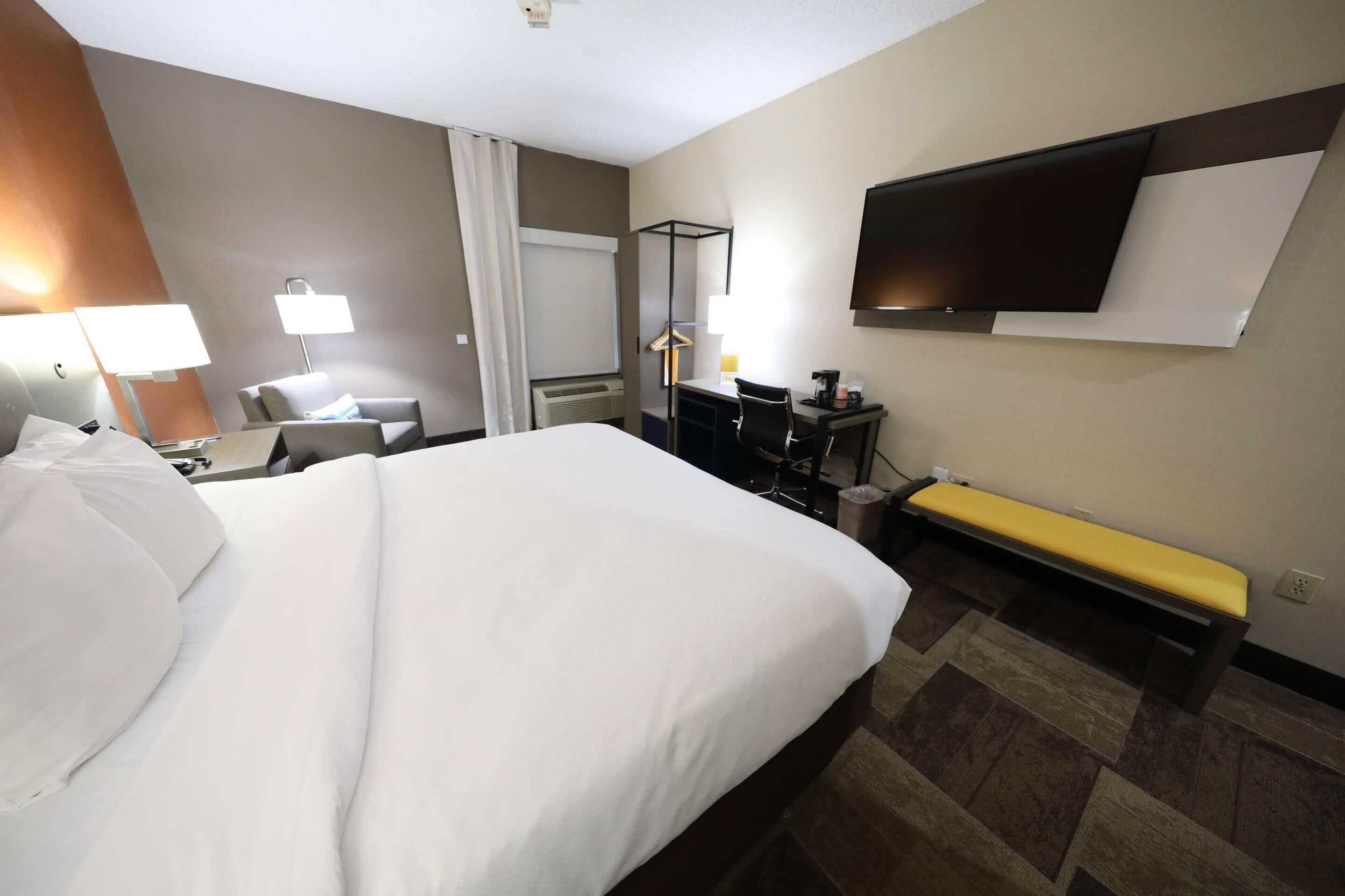 A hotel room with a large bed, a desk with a chair, a bench, a TV mounted on the wall, and a window with curtains.
