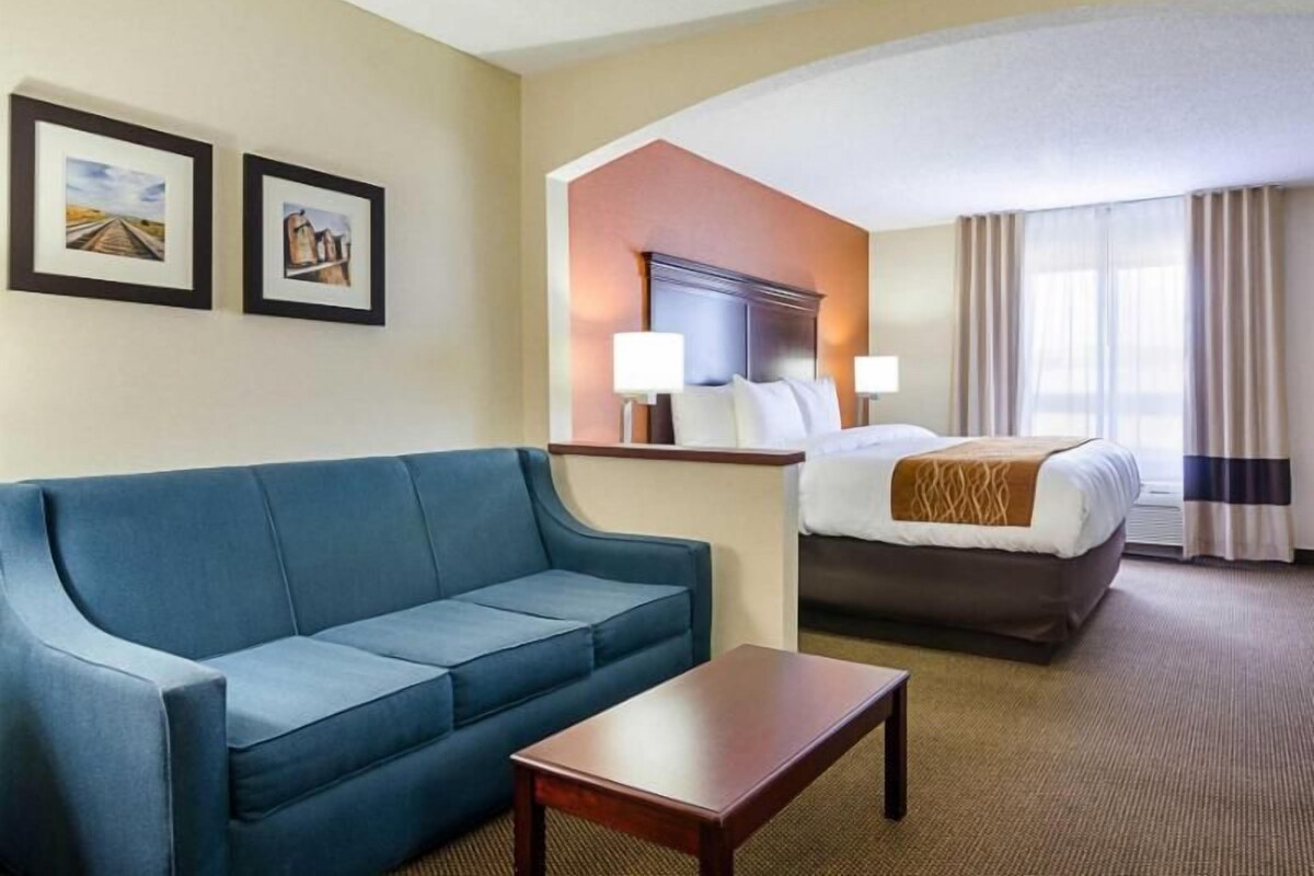 Habitación de hotel con sofá azul, mesa de madera, cama con cabecera marrón, dos lámparas fijadas a la pared y dos cuadros enmarcados.