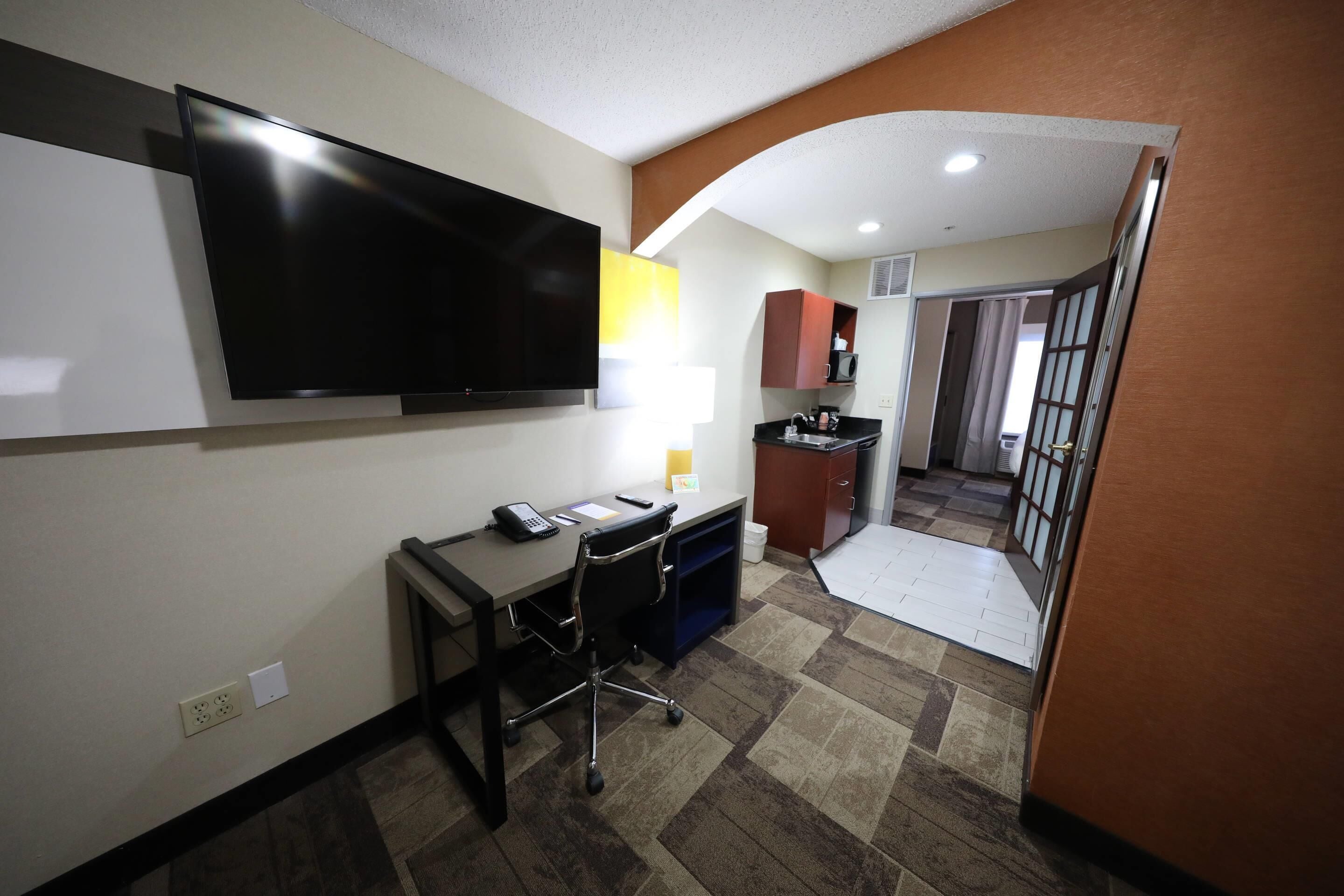 Habitación de hotel con televisión de pantalla plana, escritorio con silla, microondas y kitchenette.