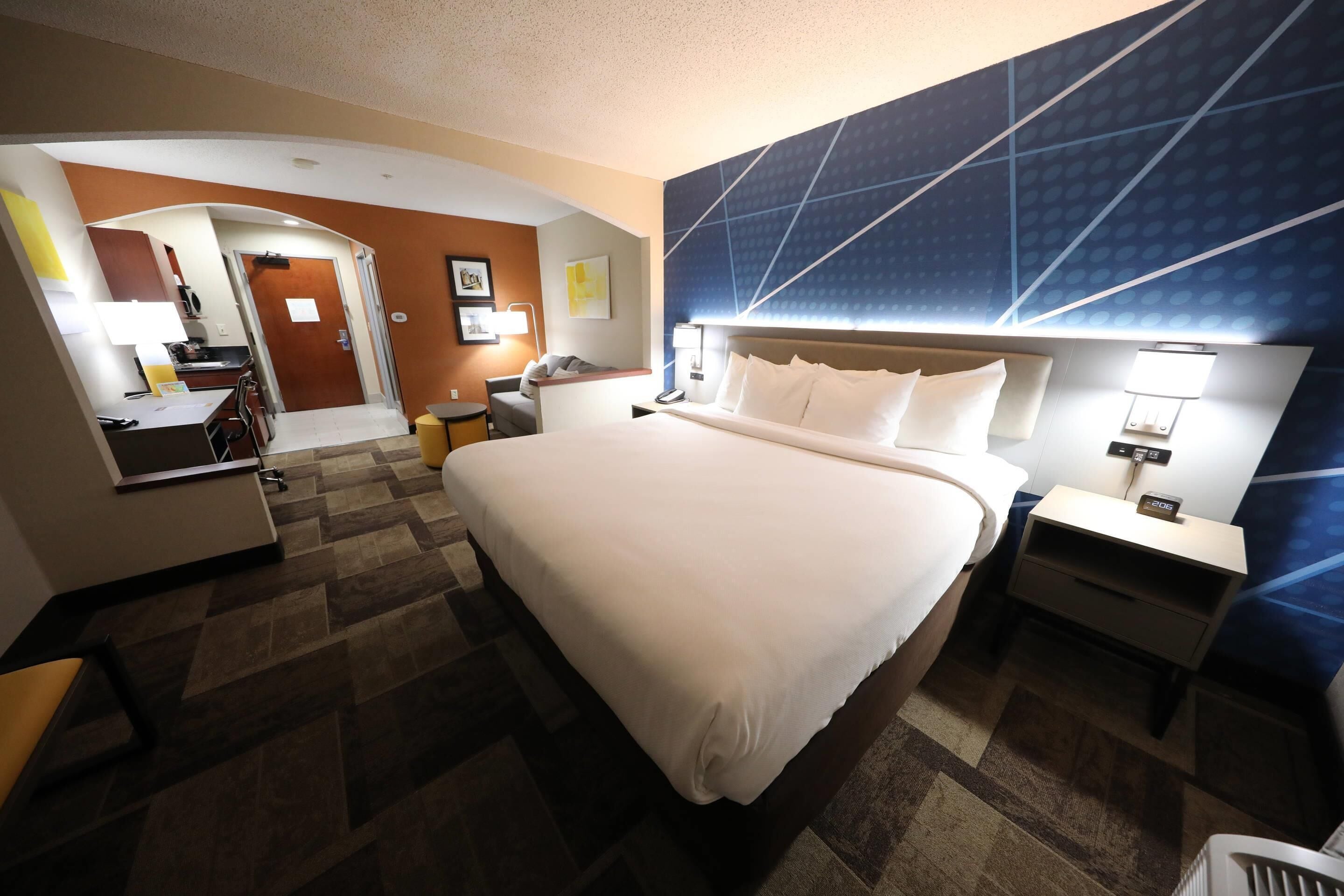 A hotel room with a large bed, a desk, a chair, a desk lamp, and a nightstand with a lamp.