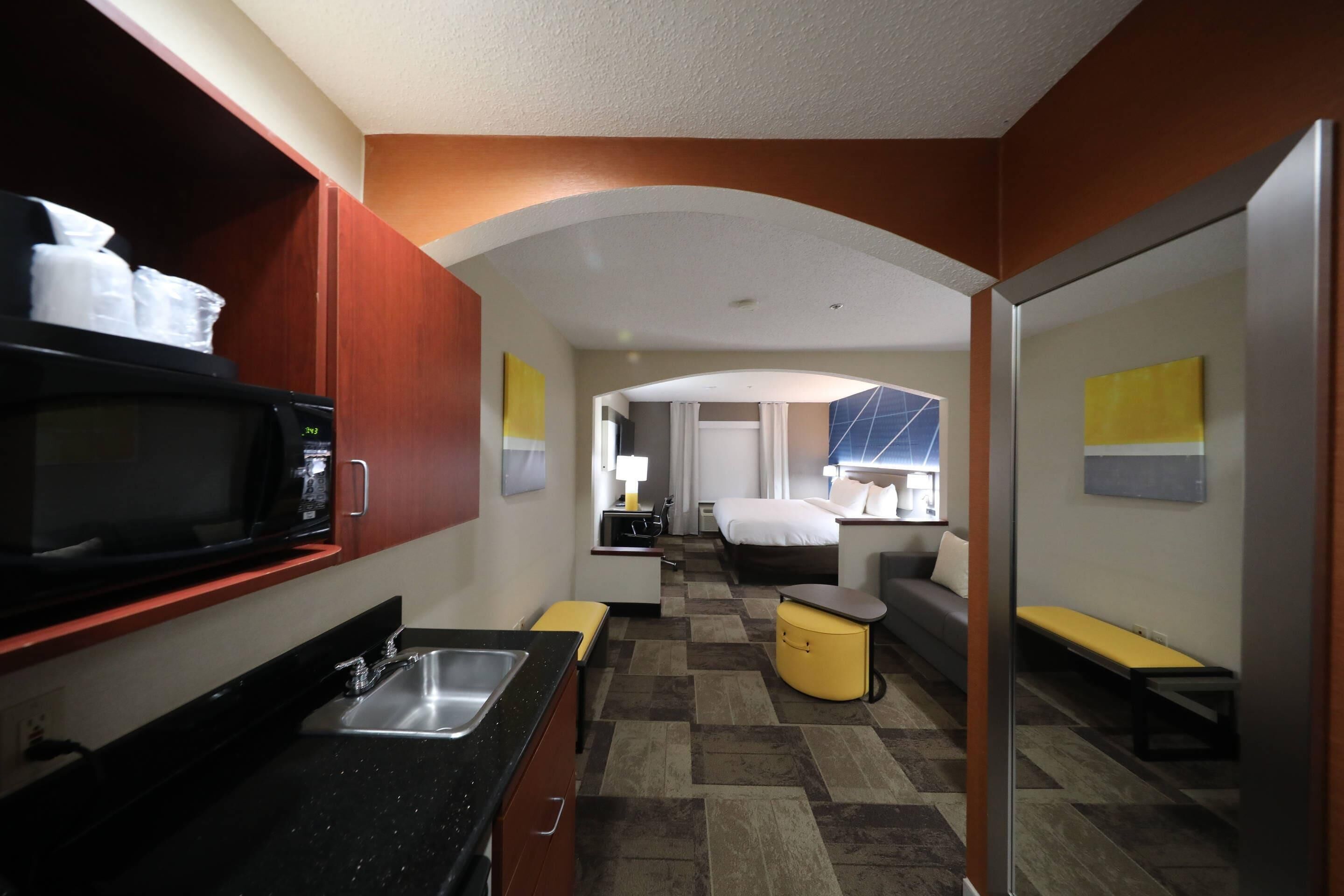 A compact hotel room with a kitchenette, a bed, a sofa, and a small seating area.
